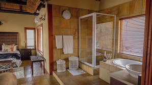 Une salle de bain spacieuse avec une grande douche, une baignoire et un lavabo. On y trouve un lit avec une tête de lit et une petite table avec une lampe.