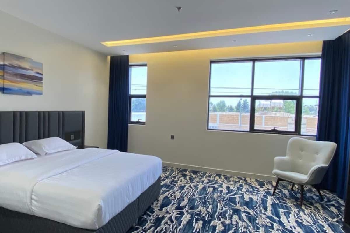 Ein Hotelzimmer mit einem Bett, einem Sessel, einem Nachttisch, einem Gemälde und großen Fenstern.