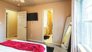 Un dormitorio con una cama, un ventilador de techo, un televisor montado en la pared y una ventana con cortinas.