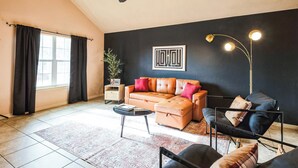Una sala de estar con un sofá, un sillón y una mesa de centro. Hay un ventilador de techo, una lámpara de pie y una ventana con cortinas.