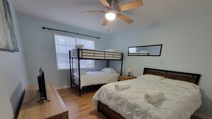 Een slaapkamer met een stapelbed, een enkel bed, een bureau met een computer, een plafondventilator en een raam met jaloezieën.