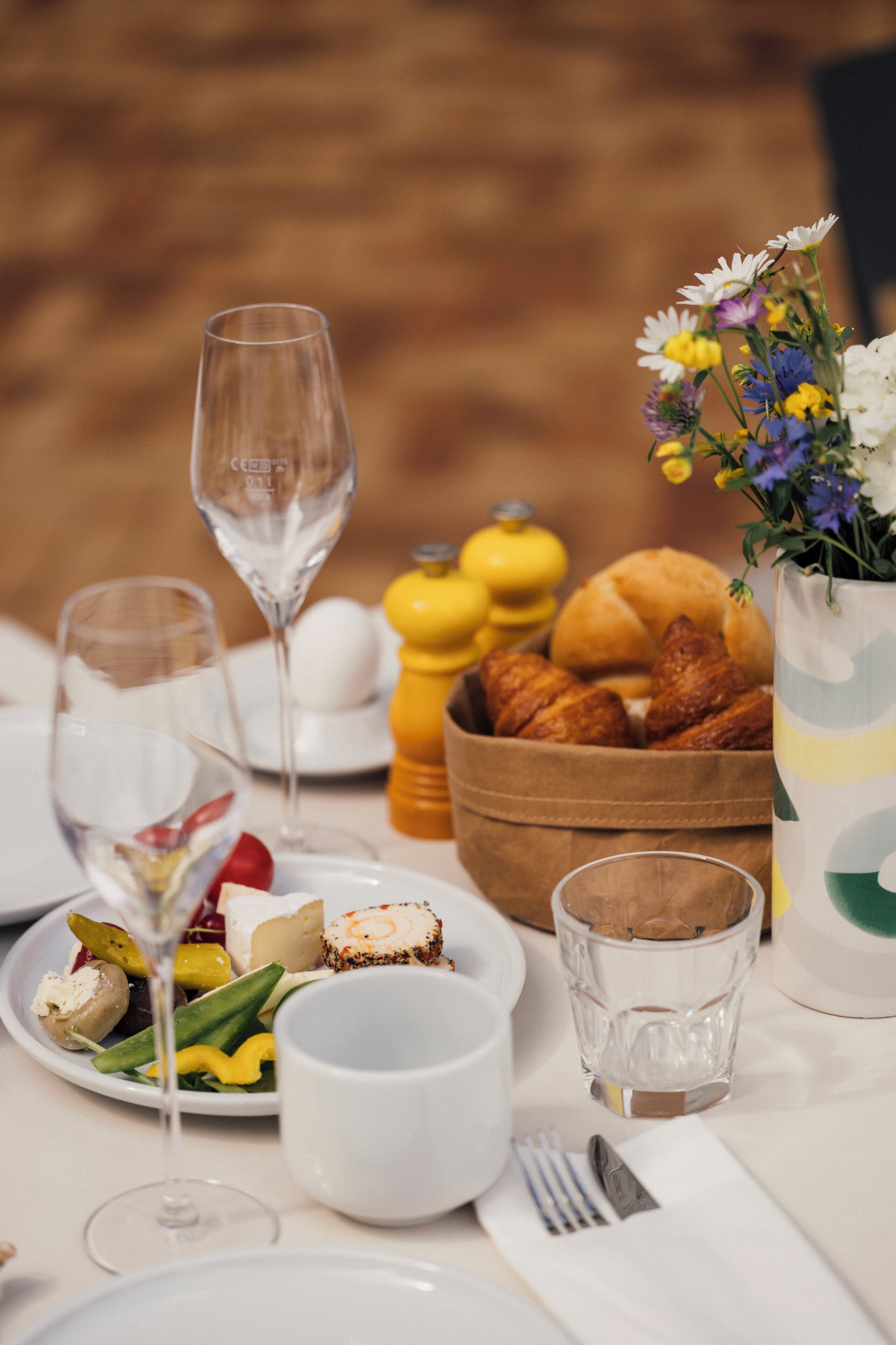 A table set with various food items, including bread, cheese, and a fruit salad, along with wine glasses and a vase of flowers.