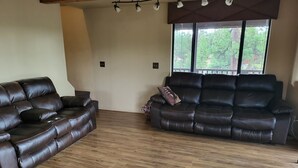 A spacious living room with two leather sofas, wooden flooring, and a large window offering a view of trees. - The Treehouse a 4-bedroom log cabin on 3 acres in Heber-Overgaard (Heber-Overgaard)
