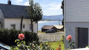 A residential area with a white house, a wooden fence, and a car parked near a body of water with mountains in the background. - 7 Person Holiday Home in Fiska (Vanylven)