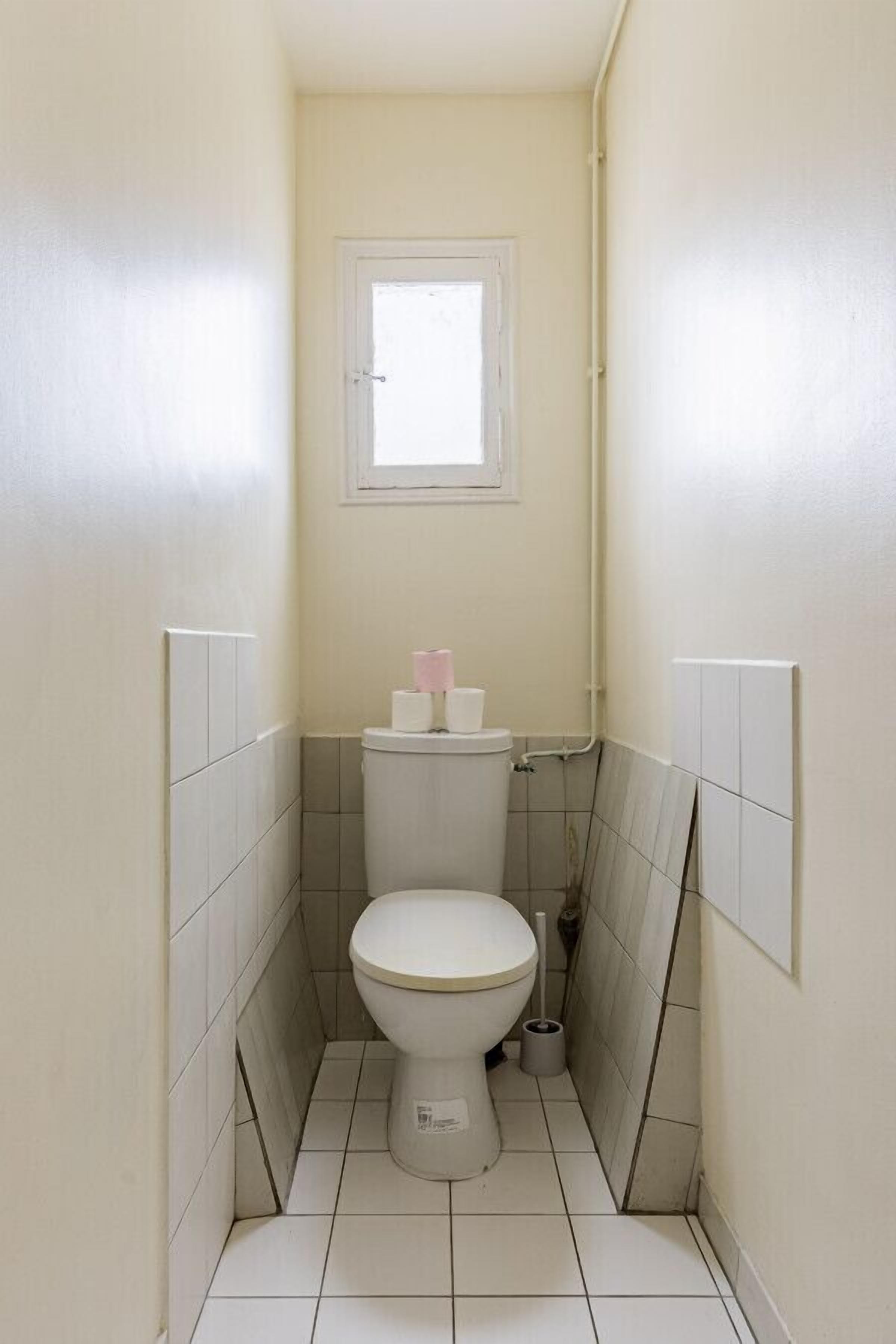 Ein kleines, schmales Badezimmer mit Toilette, einem Fenster über der Toilette und einem Toilettenpapierhalter.