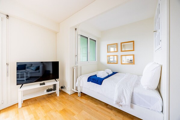A modern hotel room with a white bed, a flat-screen TV on a white stand, a radiator, and a window with blinds. - Montparnasse - Vue Tour Eiffel - Balcon (Paris)