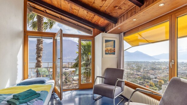 Une chambre dotée d’un lit, de deux fauteuils, d’une table et de grandes fenêtres offrant une vue sur la ville et les montagnes.