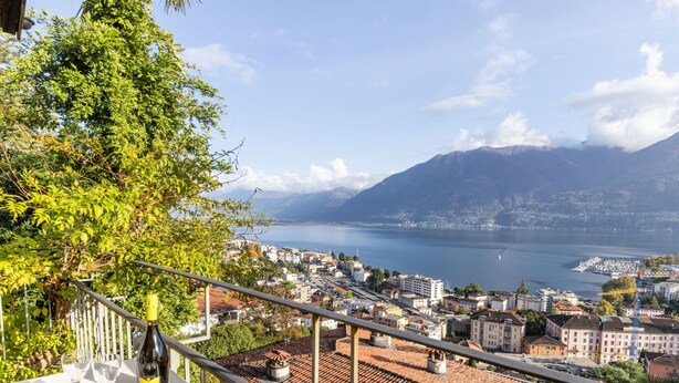 Une terrasse avec une table, une bouteille de vin et des verres, donnant sur une ville bordée d’un lac et entourée de montagnes.