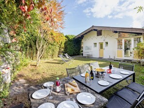 An outdoor dining area with a table set for four, surrounded by chairs. There is a small garden with a variety of plants and a tree with autumn leaves. A building with a yellow door and a glass door is visible in the background. - Egner- Apt.Caretta by Interhome (Locarno)