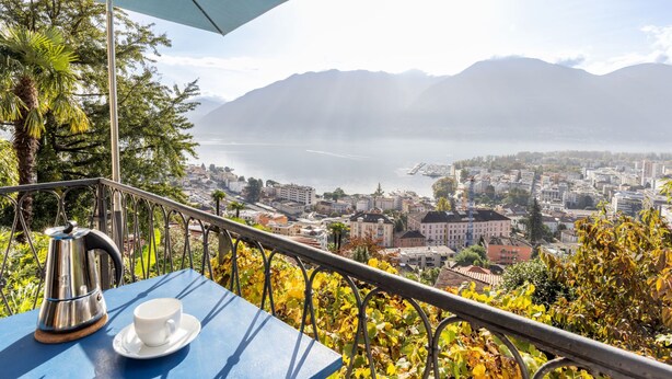 Une terrasse avec une table, une tasse et une bouilloire, d’où l’on peut admirer une ville et un lac, avec des montagnes en arrière-plan.