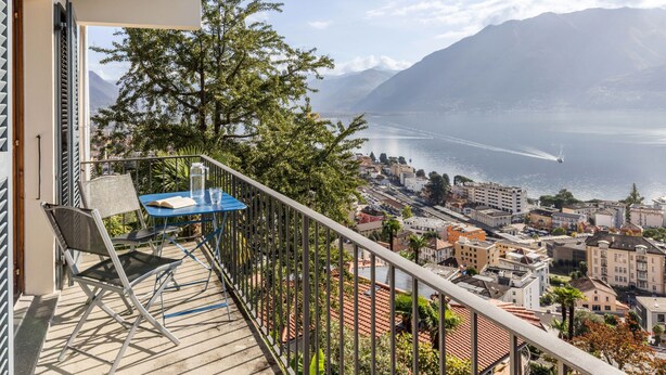 En balkon med et bord og stole, med udsigt over et bybillede og et vandområde med bjerge i baggrunden.
