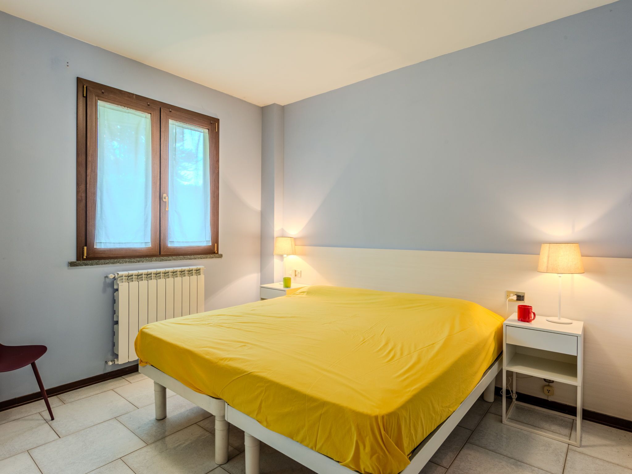 Ein Schlafzimmer mit Bett, Fenster, Heizkörper, Nachttisch, Lampe und Stuhl.