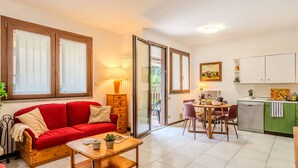 A cozy living room with a red sofa, a wooden coffee table, and a dining area with a table and chairs. There is a kitchen with white cabinets and a green countertop.