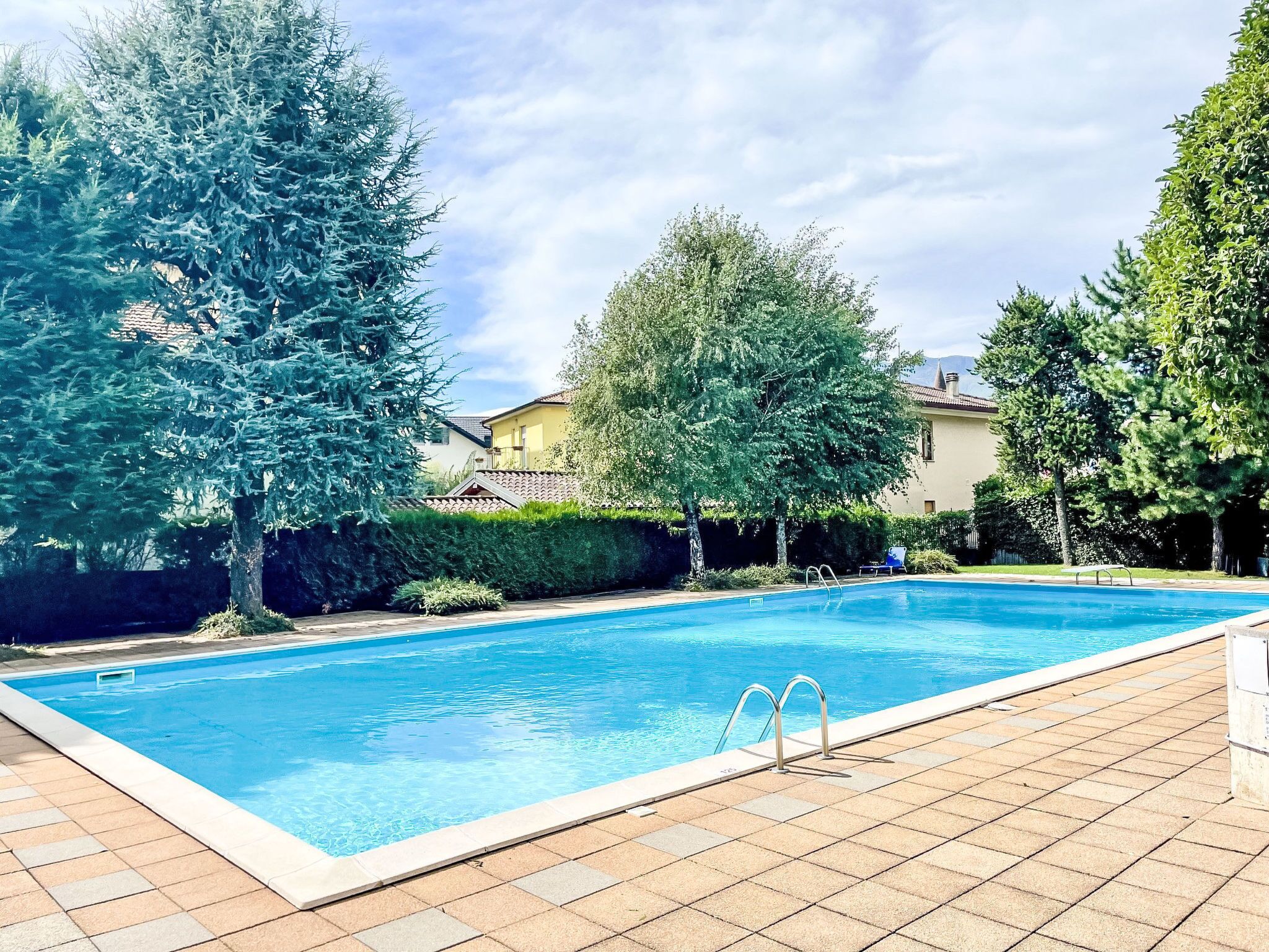 Piscina retangular com deck revestido, rodeada por árvores e um edifício ao fundo.
