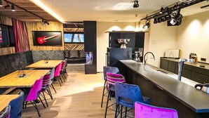 A modern dining area with wooden tables and colorful chairs, a kitchen with a bar, and a TV mounted on the wall.
