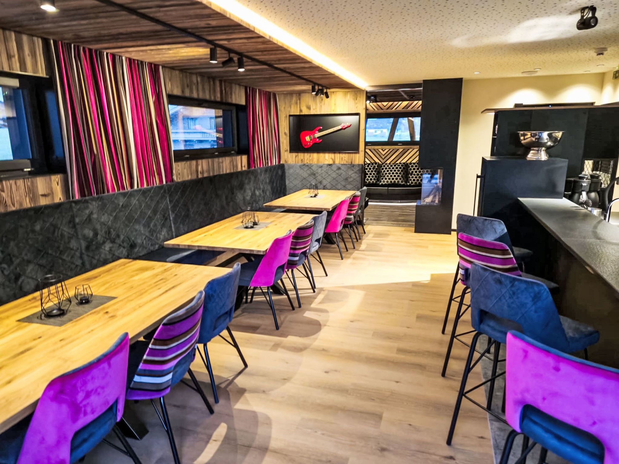 Un restaurante moderno con mesas de madera y sillas coloridas, un televisor en la pared y una zona de bar.