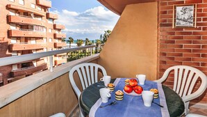 Un balcon avec une table dressée pour le petit-déjeuner, des chaises en plastique blanc et une vue sur un immeuble en briques et des palmiers.