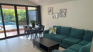 A modern living room with a green sofa, a coffee table, and a dining area with chairs and a table. There are framed pictures on the wall and a view of a pool through large windows.