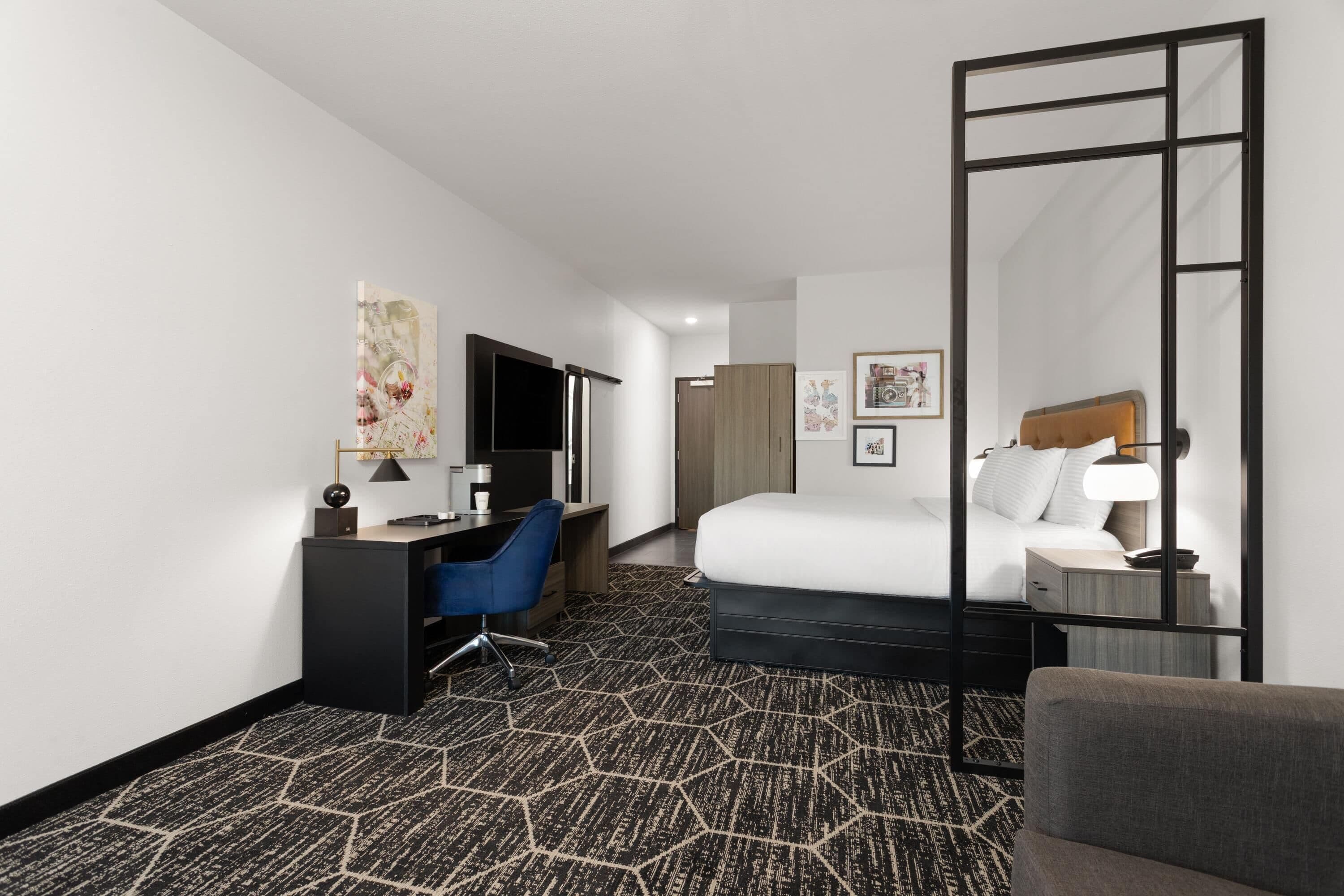 A hotel room with a bed, a desk with a chair, a large mirror, and a wardrobe.