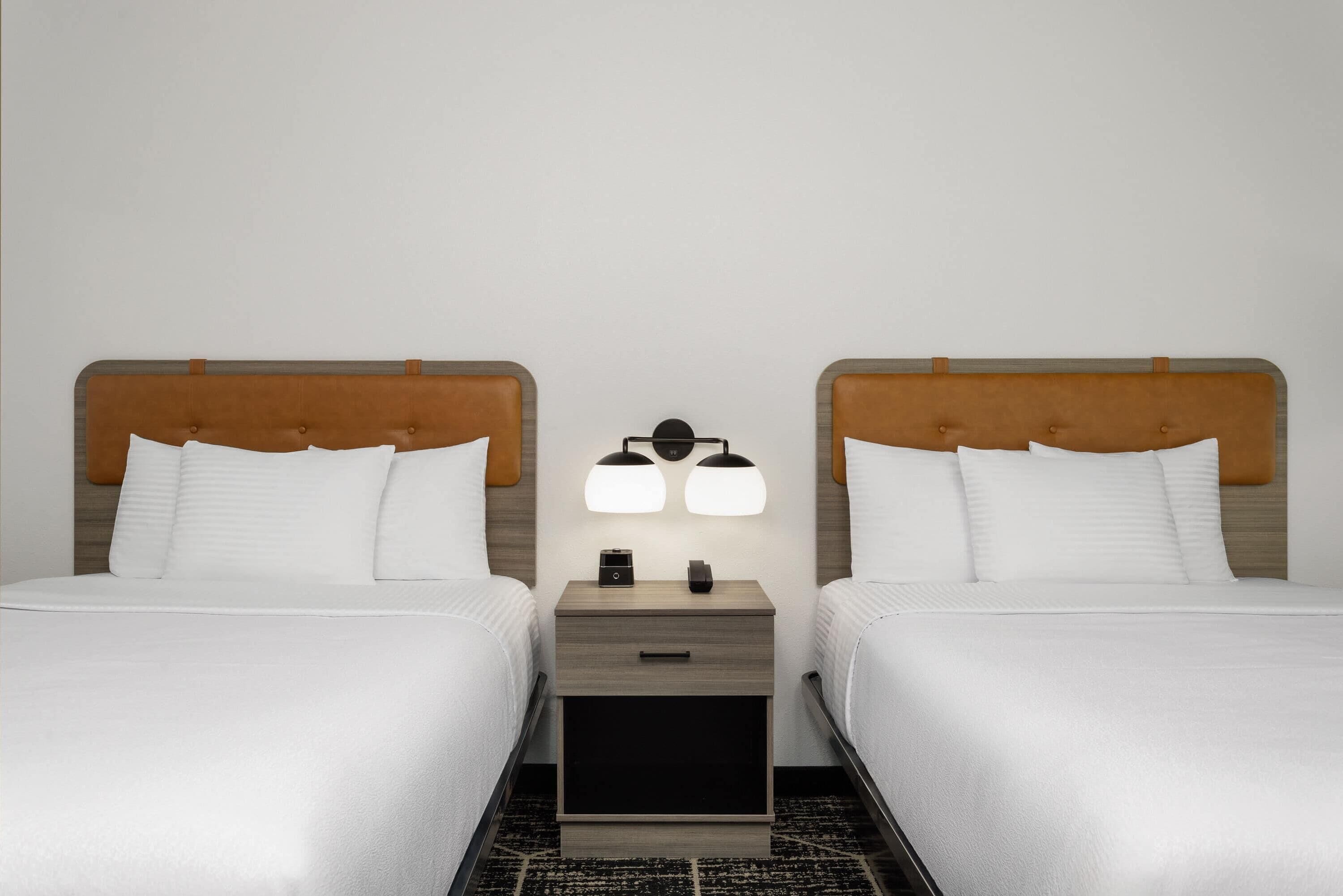 Habitación de hotel con dos camas, una mesita de noche con lámpara y un cajón pequeño.