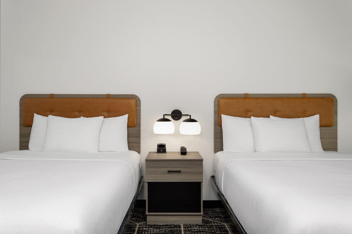 A hotel room with two beds, a nightstand with a lamp, and a small drawer.