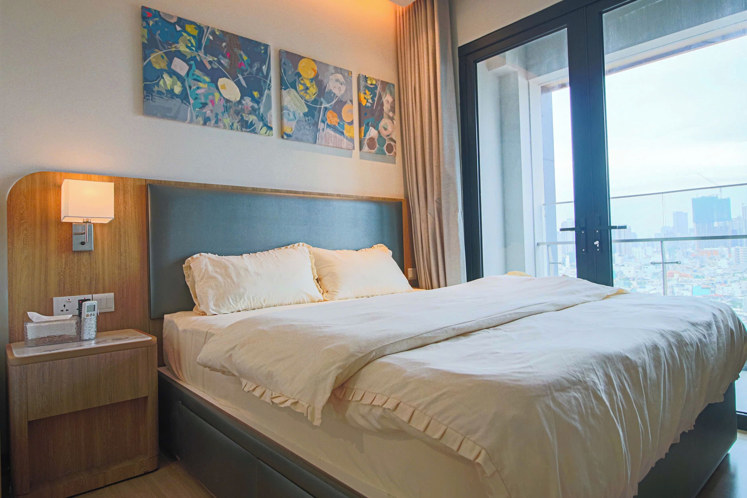 A hotel room with a large bed, a bedside table with a lamp, and a city view through a large window.