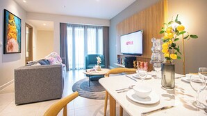 A modern living room with a grey sofa, a blue armchair, a dining table set with plates and glasses, a TV displaying 'NETFLIX', and a decorative vase with yellow flowers. - LuSSO Suites - D1mension & Zenity Tower (Ho Chi Minh City)