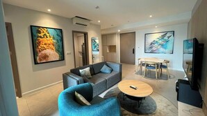 A modern living room with a grey sofa, a blue armchair, a round coffee table, and a dining area with a wooden table and chairs. - LuSSO Suites - D1mension & Zenity Tower (Ho Chi Minh City)