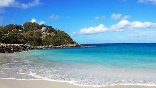 A beach with clear turquoise waters, a sandy shore, and a building on a hill with a view of the ocean.