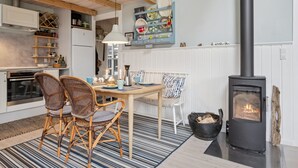 A cozy kitchen with a central dining table, wicker chairs, a black wood-burning stove, and a decorative wall shelf.