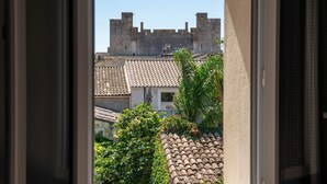 Vista a través de una ventana que muestra un edificio con tejado de baldosas, una estructura parecida a un castillo al fondo y vegetación en primer plano.