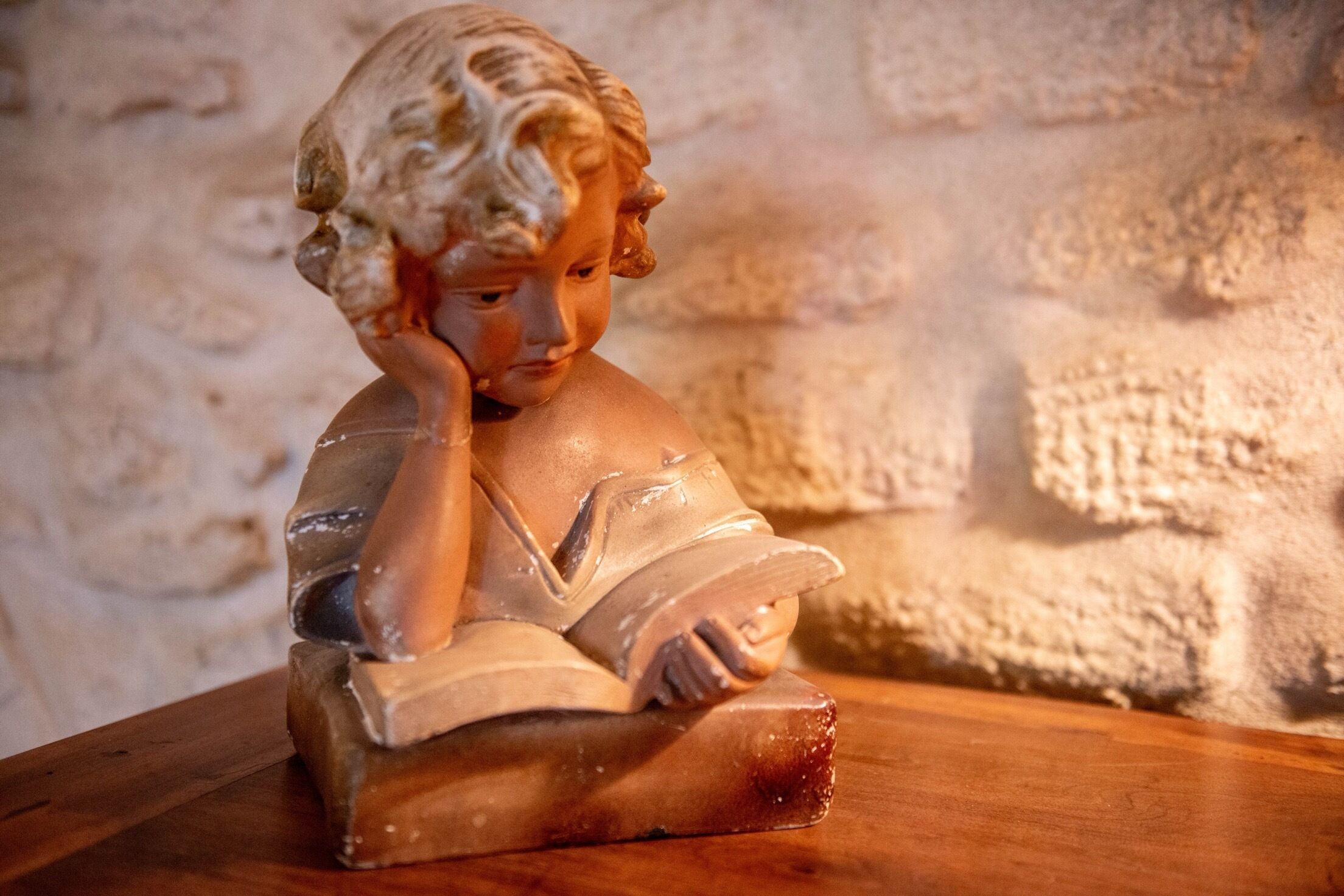 Estátua de uma criança lendo em uma mesa de madeira, com uma parede texturizada ao fundo.