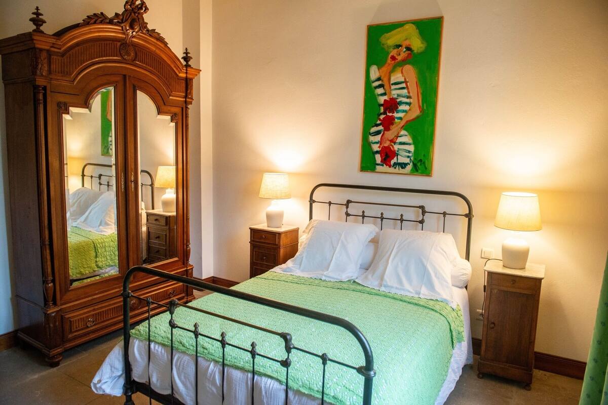 Un lit bien fait, avec du linge de lit blanc, une couverture verte et une armoire en bois dotée d’un miroir. Il y a deux tables de chevet avec des lampes.