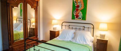 A neatly made bed with white linens, a green blanket, and a wooden wardrobe with a mirror. There are two bedside tables with lamps.