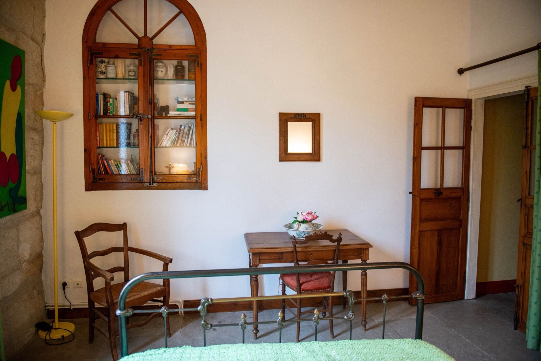 Une pièce avec une bibliothèque en bois, un bureau sur lequel se trouve un vase, une chaise, un lit avec un cadre en métal et une porte.
