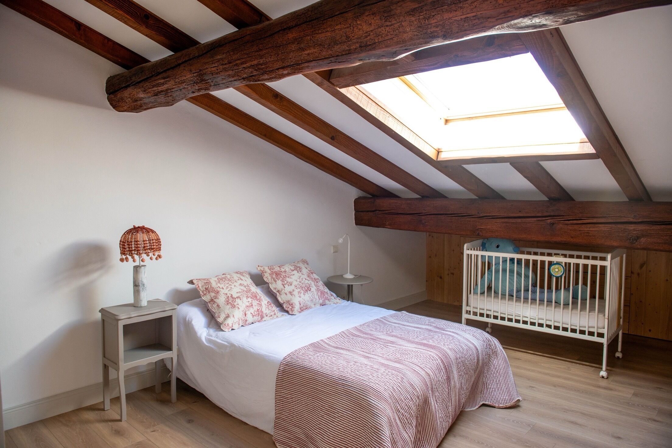 A bedroom with a sloped ceiling, a skylight, a bed with a patterned bedspread, a bedside table with a lamp, and a crib with a toy.