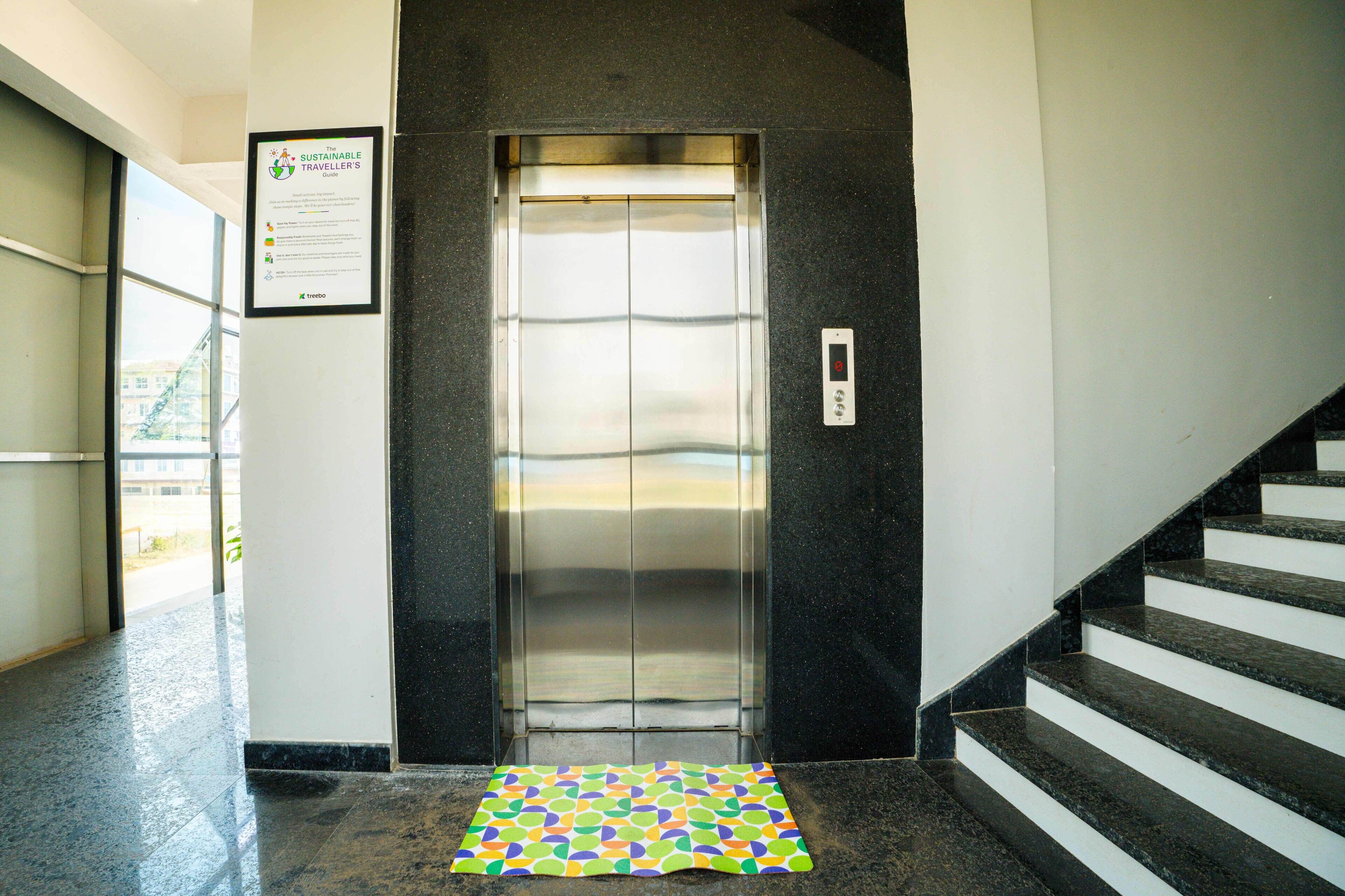An elevator with a digital control panel, a patterned mat at the entrance, and a staircase to the right.