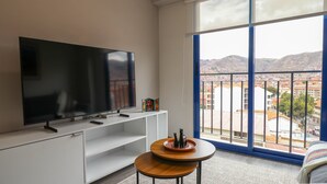 A modern living room with a large flat-screen TV, a white entertainment unit, a round wooden coffee table, and a large window with a view of a cityscape and mountains. - w* | Bright 1BR in Historic Cusco w/ Balcony (Cusco)