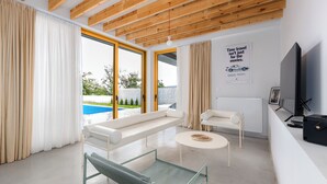 A modern living room with a glass coffee table, a sofa, and a TV. There is a large window with curtains, a pool view, and a wooden ceiling. - 3BR Luxury Villa in Tbilisi (shindisi)