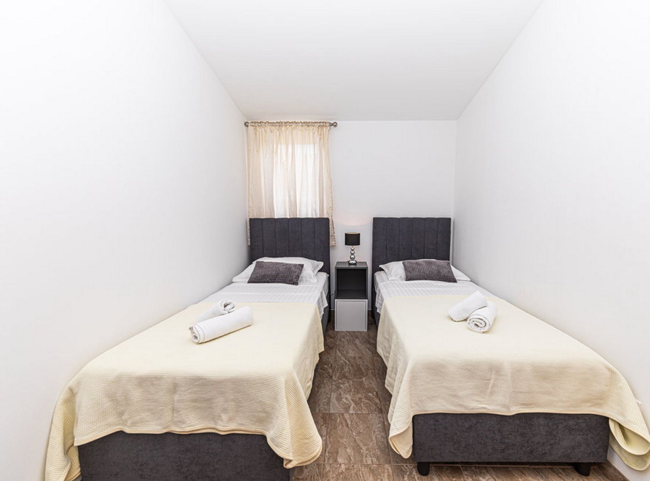 A small, clean room with two single beds, a nightstand, and a window with curtains.