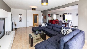 A modern living room with a sectional sofa, a glass coffee table, and a TV. There is a kitchen in the background with red cabinets and a dining area with a table and chairs. - Exquisite Spacious Villa Bufaline With Pool and Gym (Kastela)