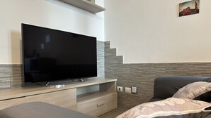 A modern bedroom with a flat-screen TV on a wooden TV stand, a grey sofa with patterned cushions, and a staircase with grey steps.