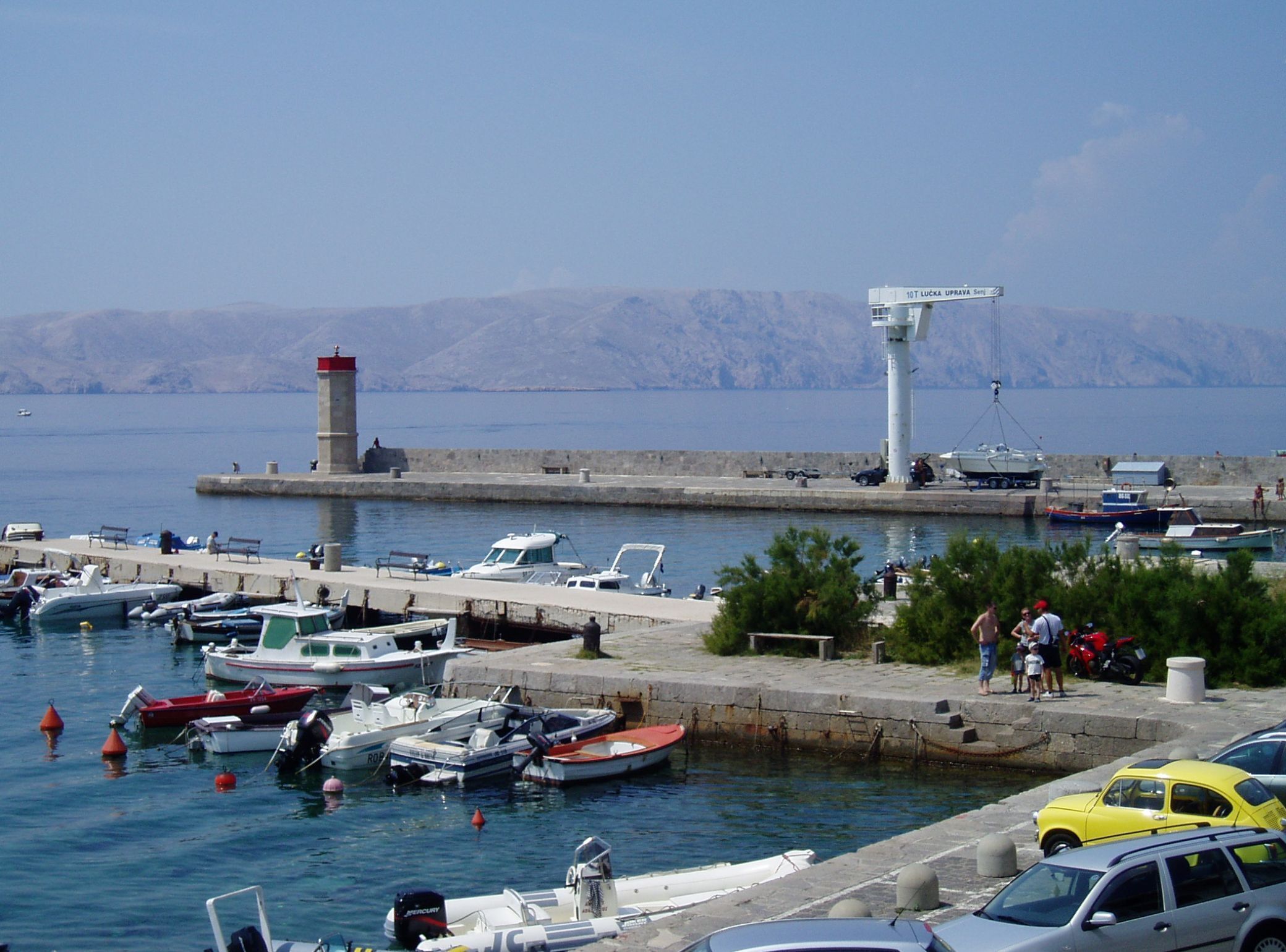 A marina with boats docked, a lighthouse, and a mountainous backdrop.