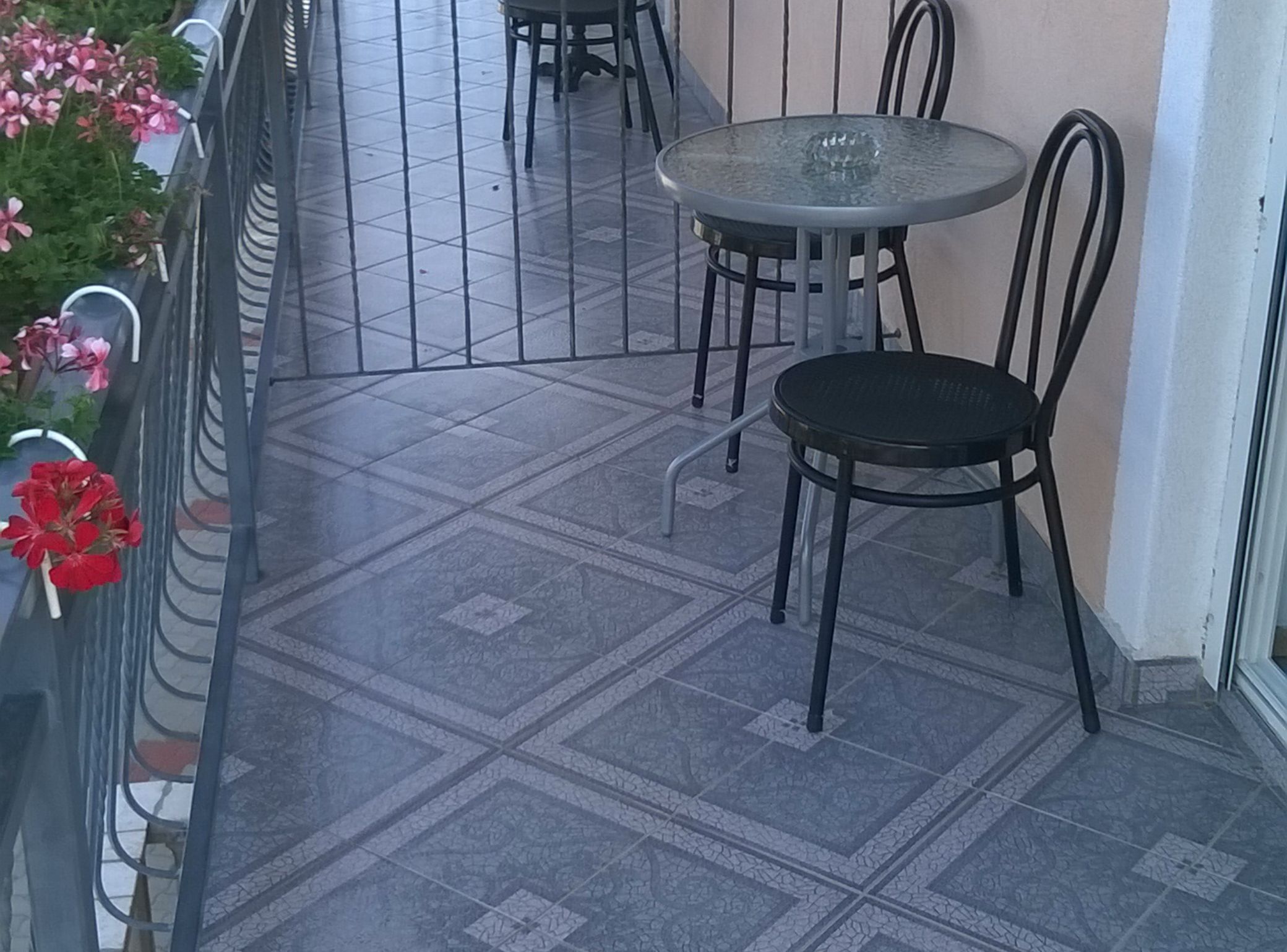 A balcony with a tiled floor, a round table with chairs, and potted plants with red flowers.