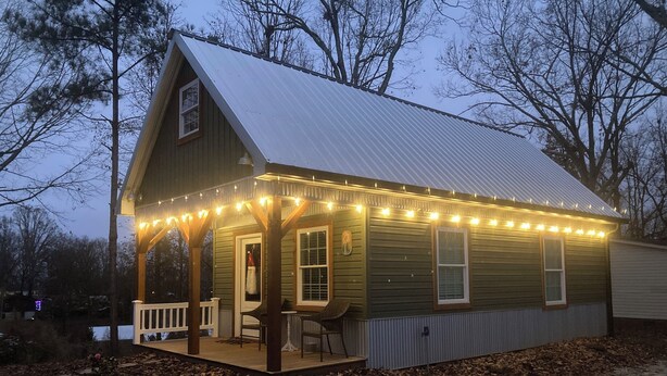 A small, single-story cabin with a porch and string lights, surrounded by trees and a dirt path. - Broyles Landing Cottage at Lake Hartwell SC (Townville)