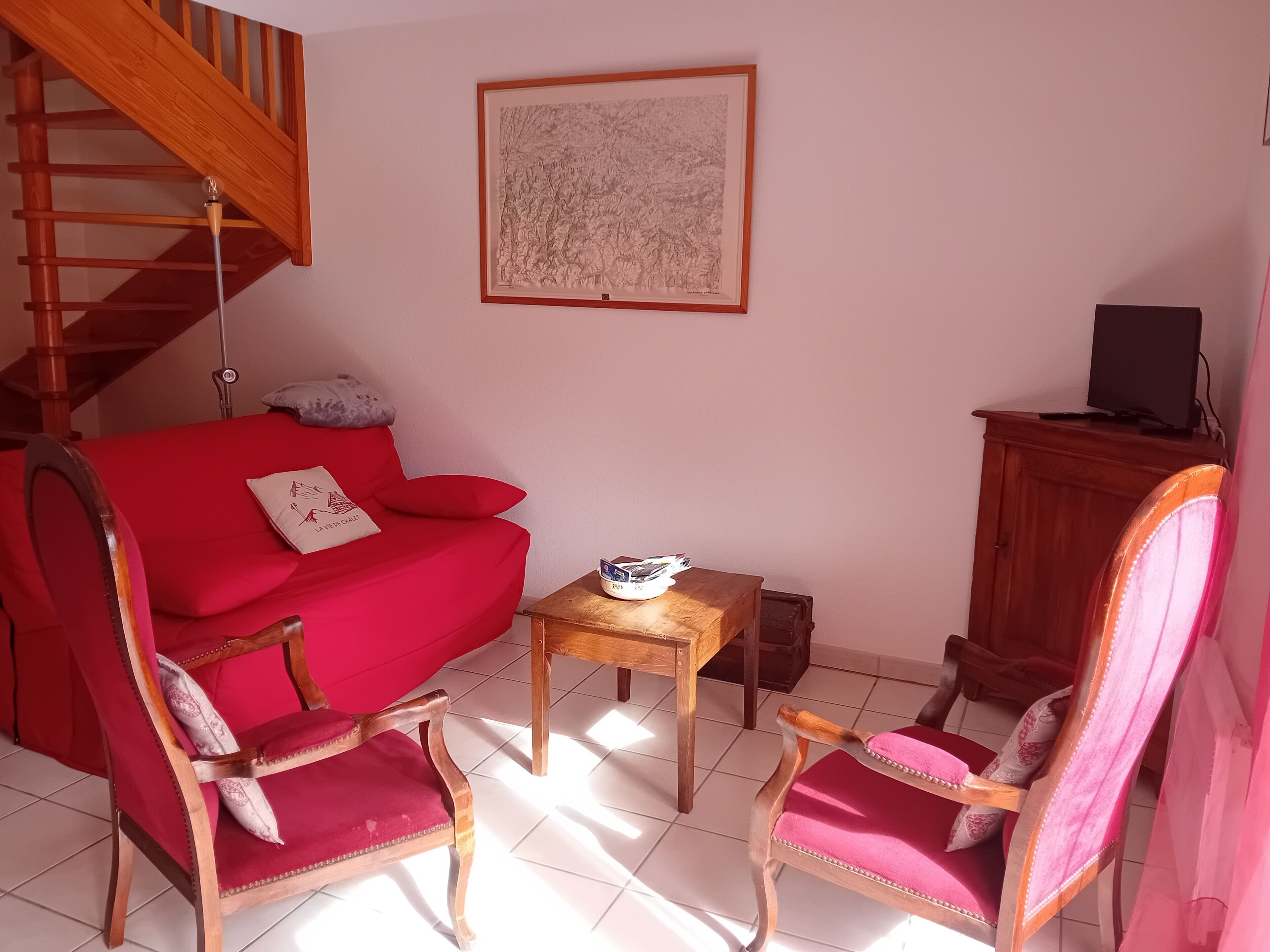 Un salon avec un canapé rouge, deux fauteuils assortis, une petite table en bois, une œuvre d’art encadrée au mur, un escalier et un téléviseur posé sur un meuble.