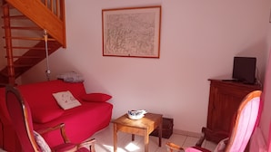 Una sala de estar con un sofá rojo, dos sillones a juego, una pequeña mesa de madera, una obra de arte enmarcada en la pared, una escalera y un televisor sobre un mueble.