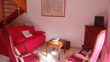 Un salon avec un canapé rouge, deux fauteuils assortis, une petite table en bois, une œuvre d’art encadrée au mur, un escalier et un téléviseur posé sur un meuble.