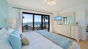 Un dormitorio con una cama grande, un aparador, televisión y un balcón con vista al océano.