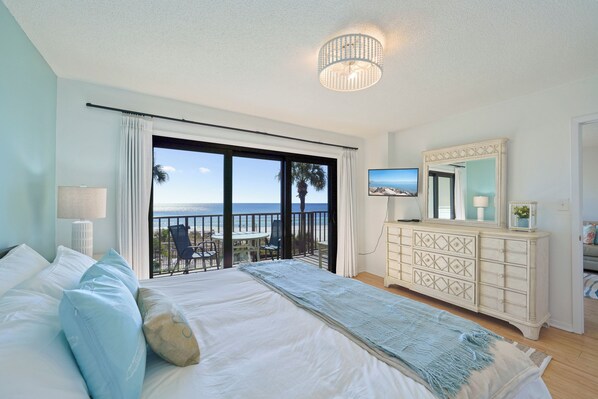 A bedroom with a large bed, a dresser, a television, and a balcony with a view of the ocean.